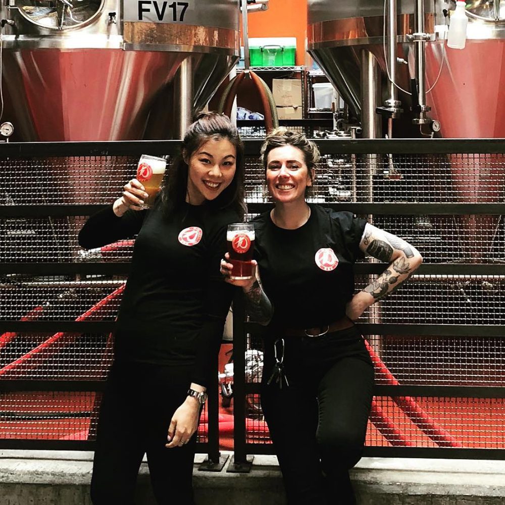 Photo of two happy young women working for HK YAU smiling at camera with their beer in front of big machines is used as HK YAU social media post